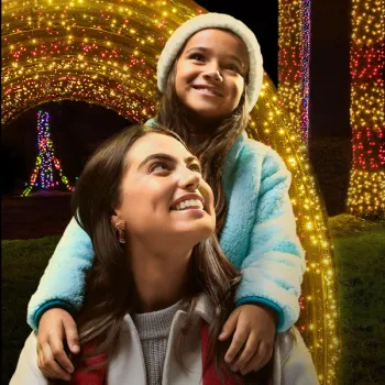 A woman with a red scarf carries a smiling girl in a blue jacket on her shoulders in front of bright holiday lights at night.