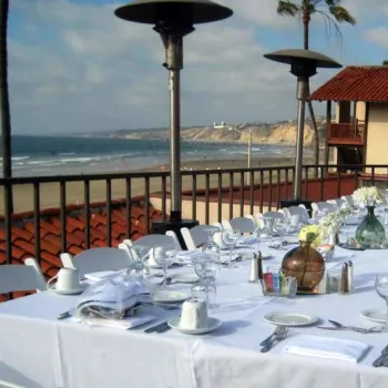 Long banquet table set for a formal meal on an outdoor terrace overlooking the beach and ocean, with white tablecloths, dishes, and decorative vases.