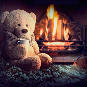 A teddy bear wearing a scarf holds a white mug while sitting in front of a lit fireplace on a furry blanket.