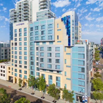 A modern mid-rise hotel building with blue and beige facade, large windows, and a vertical mural, located on a city street with trees.