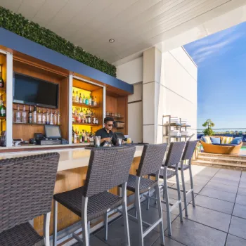 Outdoor bar with high chairs, stocked shelves of bottles, a bartender behind the counter, and a pool area with seating visible in the background under a clear sky.