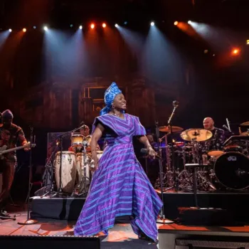 A woman in a blue and purple traditional dress sings on stage with a live band under colorful stage lights.