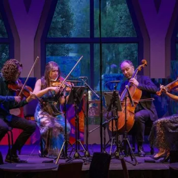 A string quartet performs onstage in front of large windows, each musician playing a different string instrument under colorful lighting.