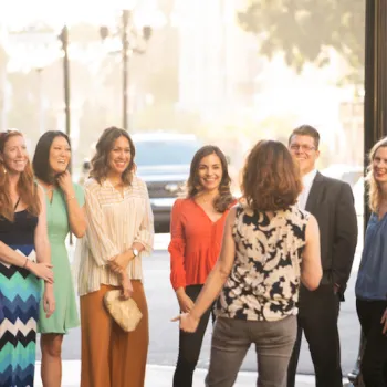 A group of eight adults standing outdoors on a city sidewalk, smiling and interacting with a woman facing them.