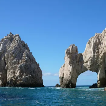 Large rock formations rise from the blue ocean under a clear sky, with one formation featuring a natural arch near the shoreline.