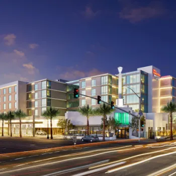 A modern hotel building at dusk with illuminated windows and palm trees along the street; light trails from passing cars are visible in the foreground.