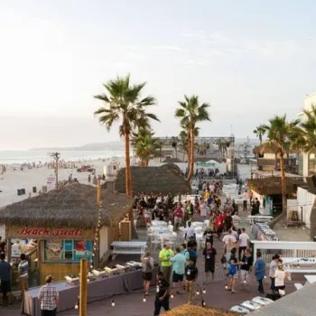 Crowds walk along a beachside boardwalk lined with palm trees, small huts, and outdoor seating; the sandy beach and ocean are visible in the background.