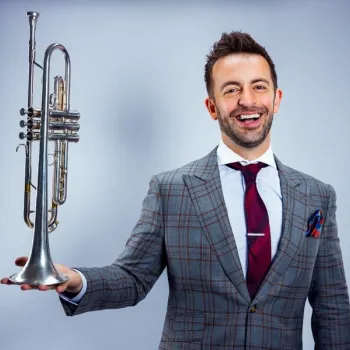 A man in a plaid suit holds a silver trumpet vertically with one hand and smiles at the camera against a plain background.
