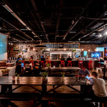 Spacious bar with wooden tables, red bar stools, industrial ceiling, and ambient lighting; people are seated and socializing.