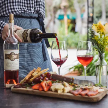 A person pours red wine into a glass next to a bottle, with a charcuterie board of bread, fruit, cheese, and meats on a table; a vase with yellow flowers is also on the table.