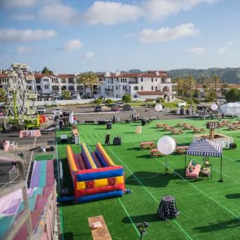 Outdoor event setup on a large green lawn with picnic tables, striped tents, inflatable games, and a Ferris wheel in the background, surrounded by buildings and hills under a blue sky.