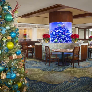 A decorated Christmas tree stands in a restaurant dining area with wooden tables and chairs, featuring a large cylindrical aquarium as a centerpiece.