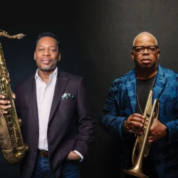 Two men pose against a dark background; one holds a saxophone, wearing a dark blazer, and the other holds a trumpet, wearing glasses and a blue patterned jacket.