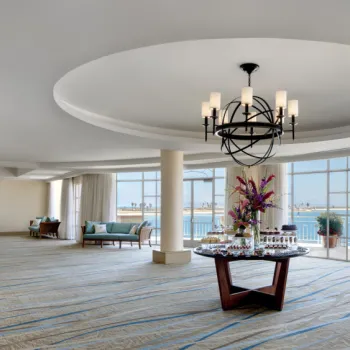 Spacious room with large windows overlooking water, a round table with flowers and drinks in the center, modern chandelier above, and seating areas by the windows.