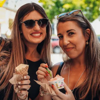 Two women smiling and holding ice cream, one in a cone and the other in a cup, outdoors on a sunny day with blurred people and cars in the background.