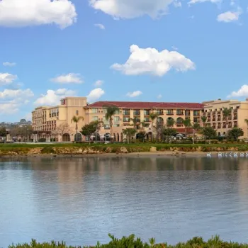 A large, multi-story waterfront building with arches and red-tiled roof sits across a calm body of water under a partly cloudy sky.