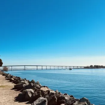 A long bridge spans across a calm body of water, with rocks and a dirt path along the shoreline under a clear blue sky.