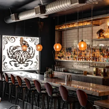 A modern bar with a marble counter, high stools, hanging pendant lights, liquor bottles on shelves, and a chalkboard menu on the wall.