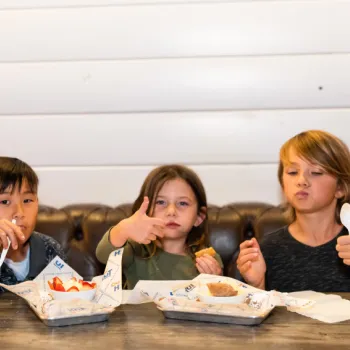Three children sit at a table eating dessert with plastic spoons, giving thumbs up and playful gestures, against a white paneled wall background.