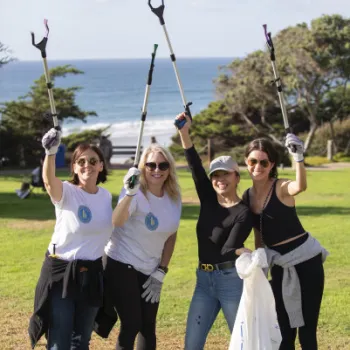 Four women pose outdoors near the ocean, holding litter grabbers and smiling, participating in a park cleanup event on a sunny day.