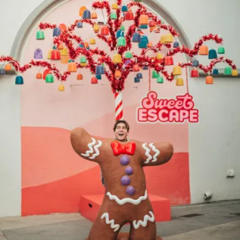 A person poses with their face in a large gingerbread man cutout, standing under a colorful, candy-themed tree with a "Sweet Escape" sign in the background.