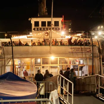 A crowd stands on the upper deck of the historic ferryboat Berkeley at night, docked at the Maritime Museum of San Diego, with lights illuminating the scene.