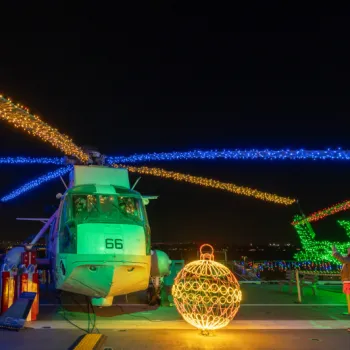 Helicopters decorated with colorful holiday lights on a ship deck at night, with people observing and a large illuminated ornament in the foreground.