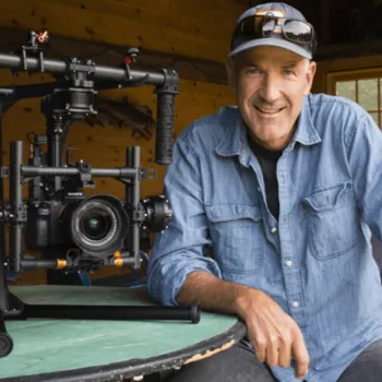 A man in a denim shirt and cap sits at a table with a professional camera stabilizer and camera in front of him inside a wooden room.