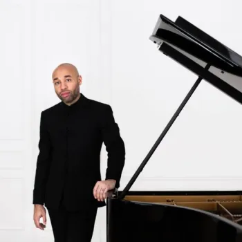 A man in a black suit stands beside a grand piano in a white, minimalist room.