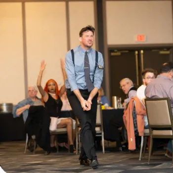 A man in a blue shirt and tie stands and gestures animatedly in front of seated people at a formal indoor event.