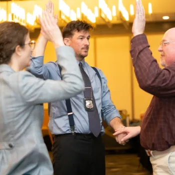 Three people stand indoors with their right hands raised; one man wears a badge on a lanyard and holds the hand of another man during what appears to be a formal or official ceremony.