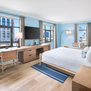 Bright hotel room with a large bed, desk, TV, sofa, and multiple windows offering a city view. Decor features light wood furniture and blue accents.