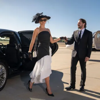A woman in a black and white dress exits a luxury car with help from a man in a suit; a private jet is visible in the background on a sunny day.