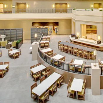 Spacious indoor dining area with long tables, wooden chairs, modern lighting, and a central bar counter in a multi-level building.