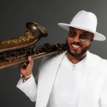 Man in a white suit and hat holds a decorated saxophone on his shoulder, smiling, against a plain dark background.