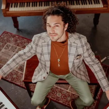 A man in a plaid blazer and green pants sits at a piano bench, hands on two keyboards, with another piano in the background on patterned rugs.