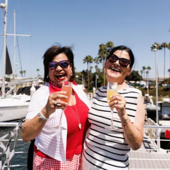 Two women smiling and holding drinks stand on a boat deck, with sailboats and palm trees in the background on a sunny day.