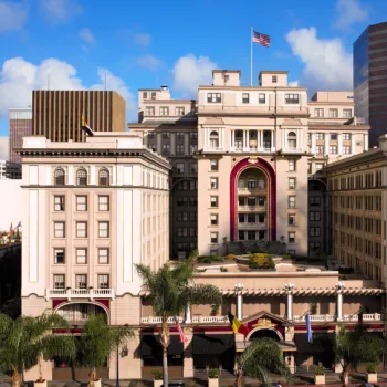 A grand historic hotel with columns and arches stands in a cityscape, flanked by palm trees and modern office buildings, with an American flag flying on top.