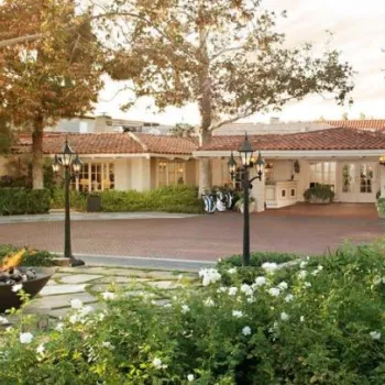 A single-story building with a red-tiled roof is surrounded by trees, green landscaping, white flowers, a cobblestone driveway, and a fountain.