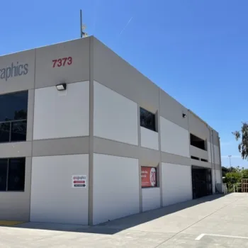 A commercial building with “alphagraphics” signage at address 7373, featuring large windows and a driveway on a clear, sunny day.