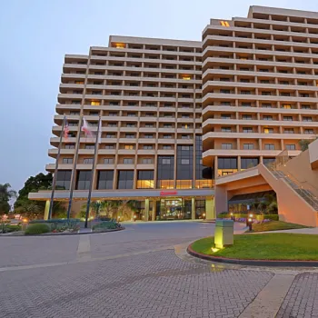 Multi-story hotel building with numerous balconies, exterior lights on, and landscaped entrance with stairs, flags, and a paved driveway in the foreground.