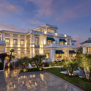 Large, elegant mansion with multiple stories, illuminated at dusk, surrounded by palm trees, a pool, and a reflective wet patio area.