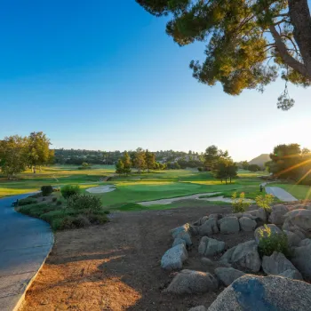 A golf course in the early morning, with sunlight streaming through trees, a paved path, sand traps, and grassy fairways in view.
