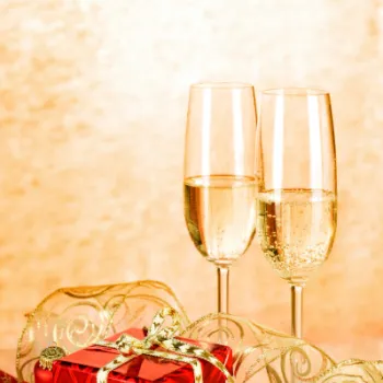 Two filled champagne flutes next to a red gift box with gold ribbon, gold ornaments, and festive decorations on a golden background.