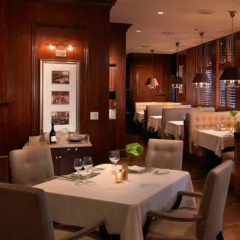 Elegant restaurant interior with wooden paneling, white tablecloths, cushioned booths, wine glasses, and soft lighting. Tables are set and a framed art piece hangs on the wall.