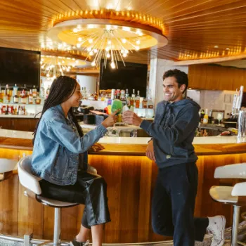 Two people sit at a bar, smiling and clinking drinks, with bottles and a decorative light fixture in the background.