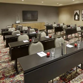 Classroom-style meeting room with rows of tables and chairs, notepads, glasses, and water pitchers, set on a patterned carpet with framed photos on the wall.