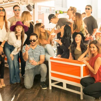 A group of people pose and smile together at an outdoor gathering, some sitting and others standing, with drinks in hand on a sunny day.