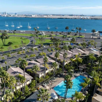 Aerial view of a waterfront area with a marina, sailboats, palm trees, buildings, a swimming pool, and a city skyline in the distance under a clear sky.
