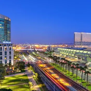 Cityscape at dusk showing the Hilton hotel, palm trees, roads with light trails, and a modern convention center near a waterfront.